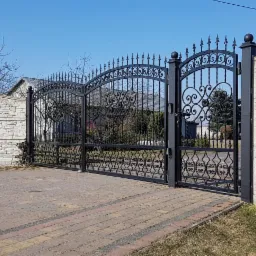 Czarna, dwuskrzydłowa brama wjazdowa z kutymi zdobieniami, zamontowana między dwoma murowanymi filarami z jasnego kamienia. Brukowana droga prowadzi w kierunku bramy.