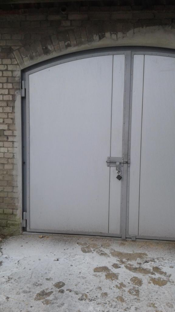 Szara, dwuskrzydłowa brama garażowa z zamkiem na tle ceglanego muru z łukowatym nadprożem.