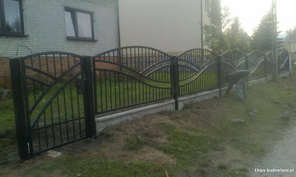 Czarna, metalowa brama i ogrodzenie z falistym wzorem, osadzone na betonowej podmurówce, widoczne na tle domu z cegły i trawnika, z osobą pracującą przy ziemi w tle.