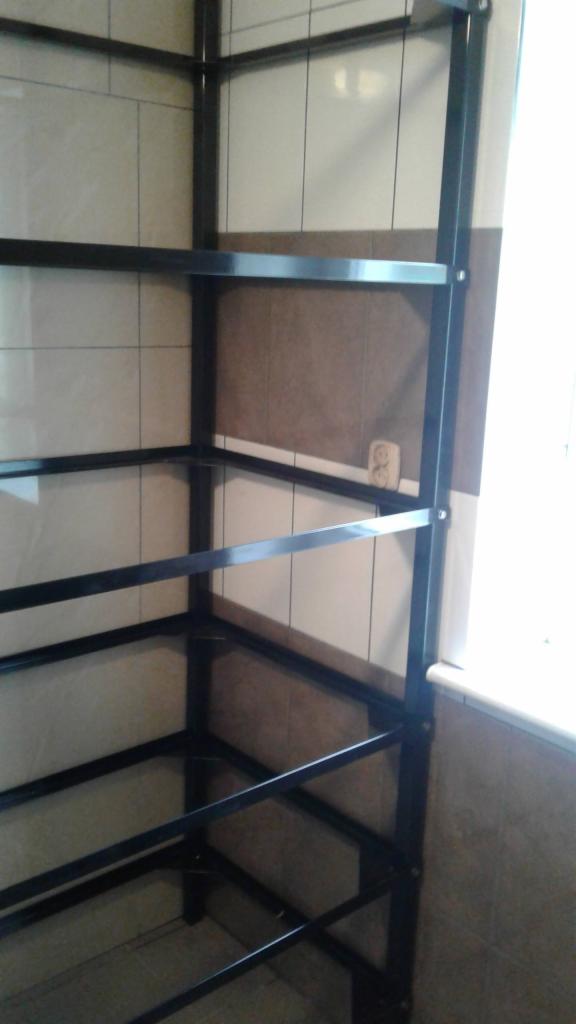 Czarna, metalowa konstrukcja regału z półkami, zamontowana w rogu pomieszczenia z wyłożonymi płytkami ścianami, widoczne śruby mocujące.