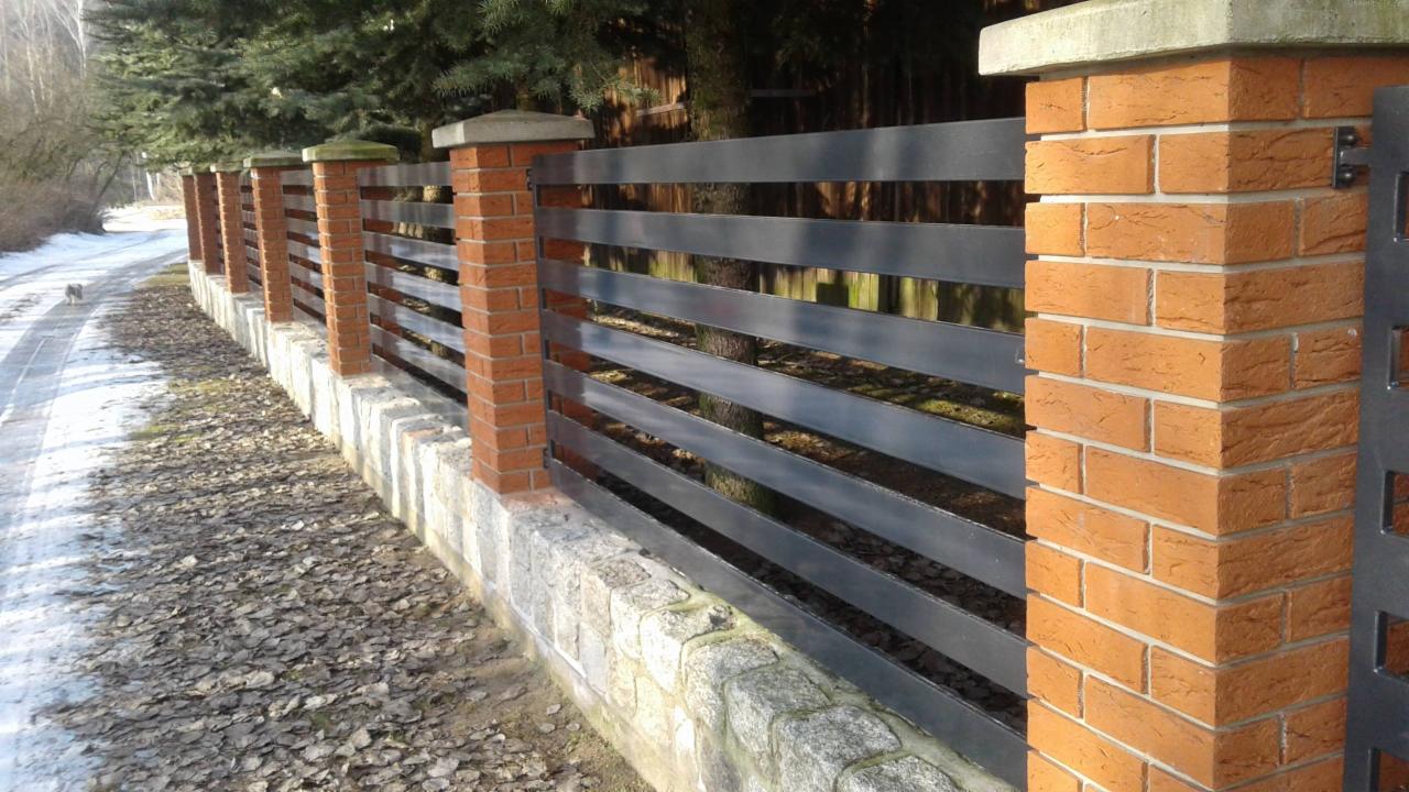 Nowoczesne ogrodzenie z poziomych, grafitowych profili metalowych, osadzone na niskim murku z kamienia polnego, z ceglanymi słupkami zwieńczonymi betonowymi czapami, na tle zimowego krajobrazu.
