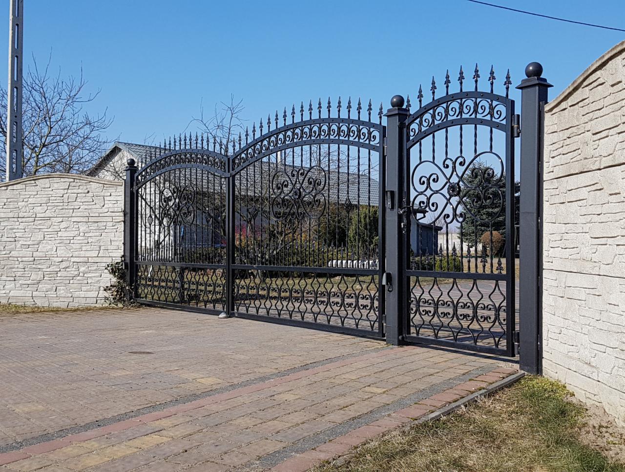 Czarna, dwuskrzydłowa brama wjazdowa z kutymi zdobieniami, zamontowana między dwoma murowanymi filarami z jasnego kamienia. Brukowana droga prowadzi w kierunku bramy.