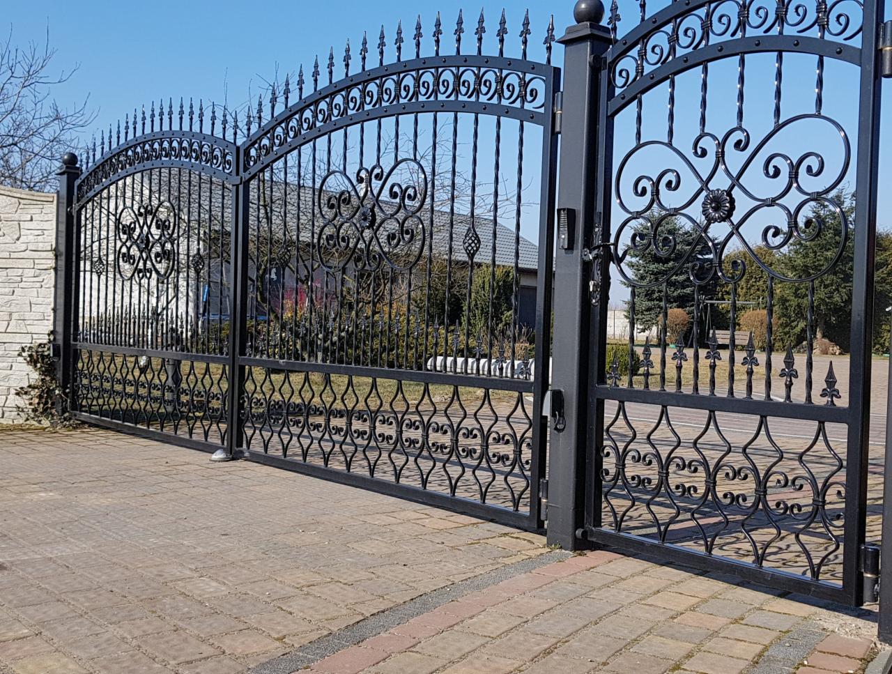 Czarna, dwuskrzydłowa brama wjazdowa z kutymi, ozdobnymi elementami, widoczna z perspektywy chodnika z kostki brukowej na tle domu i ogrodu.