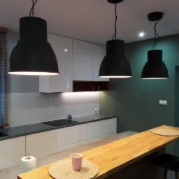 Nowoczesna kuchnia z białymi szafkami, szarym blatem, drewnianym barkiem i trzema czarnymi lampami wiszącymi w stylu industrialnym.