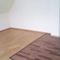 Podłoga w pokoju z dwoma rodzajami wykończenia: jasne panele imitujące drewno i ciemniejsze płytki ułożone w cegiełkę, oddzielone czarną listwą.