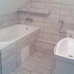 Wykończona łazienka w szarych płytkach imitujących drewno, z wanną obudowaną płytkami, umywalką z szafką i półką nad wanną.