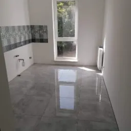 Wnętrze łazienki w trakcie prac wykończeniowych: położone szare płytki podłogowe, fragment ściany z dekoracyjnymi płytkami nad instalacją wodną, białe ściany i okno z widokiem na zieleń.