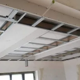 Konstrukcja sufitu podwieszanego z metalowych profili i częściowo zamontowanych płyt gipsowo-kartonowych, widoczny fragment instalacji wentylacyjnej i surowy stan ścian.