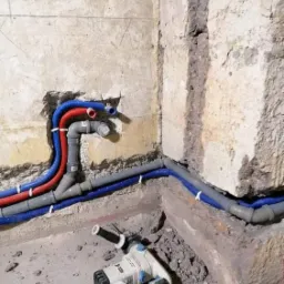 Wykute w starej ścianie bruzdy z nową instalacją wodną z rur w kolorach czerwonym i niebieskim, przymocowanych białymi uchwytami, obok leży zakurzona wiertarka udarowa.