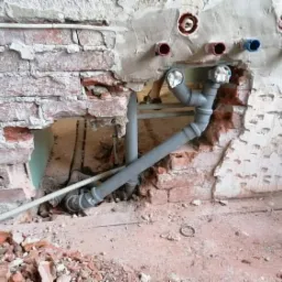 Odsłonięta instalacja wodna w trakcie prac remontowych, widoczne rury PCV, fragment muru z cegły i gruz na podłodze, przygotowane otwory na przyłącza.