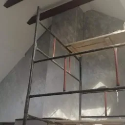 Pracownik na rusztowaniu maluje ścianę w szarym odcieniu w pokoju na poddaszu z oknem dachowym, widoczna faktura ściany.