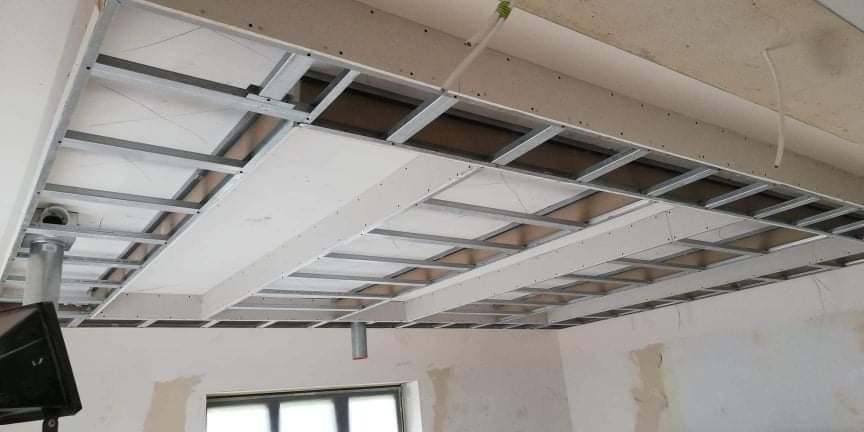 Konstrukcja sufitu podwieszanego z metalowych profili i częściowo zamontowanych płyt gipsowo-kartonowych, widoczny fragment instalacji wentylacyjnej i surowy stan ścian.