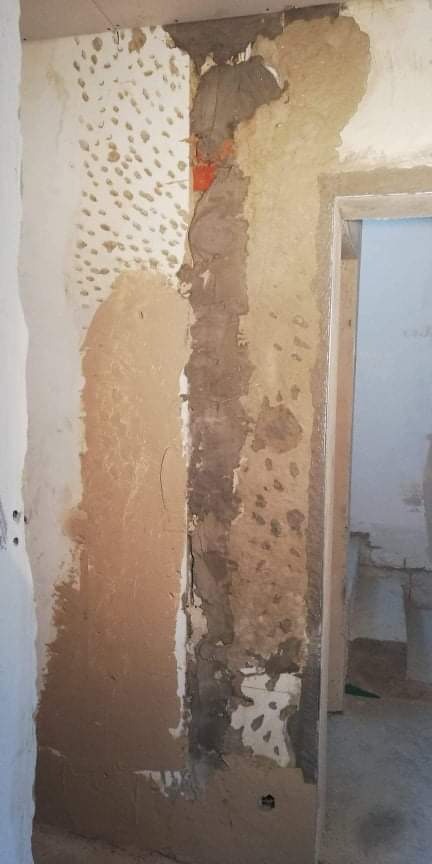 Przygotowanie ściany do dalszych prac: warstwy starego tynku z widocznymi śladami po wcześniejszych naprawach, częściowo usunięte, odsłaniające fragment cegły, obok ościeżnicy drzwiowej.