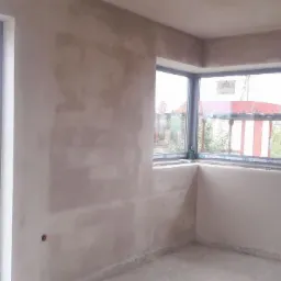 Pokój w stanie deweloperskim z nałożonymi tynkami maszynowymi, widoczne dwa okna z szarymi ramami, przez jedno z okien widać zieleń i czerwony samochód.