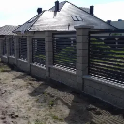 Nowoczesne ogrodzenie posesji z betonowych słupków i poziomych, metalowych przęseł, widoczne z perspektywy drogi gruntowej.