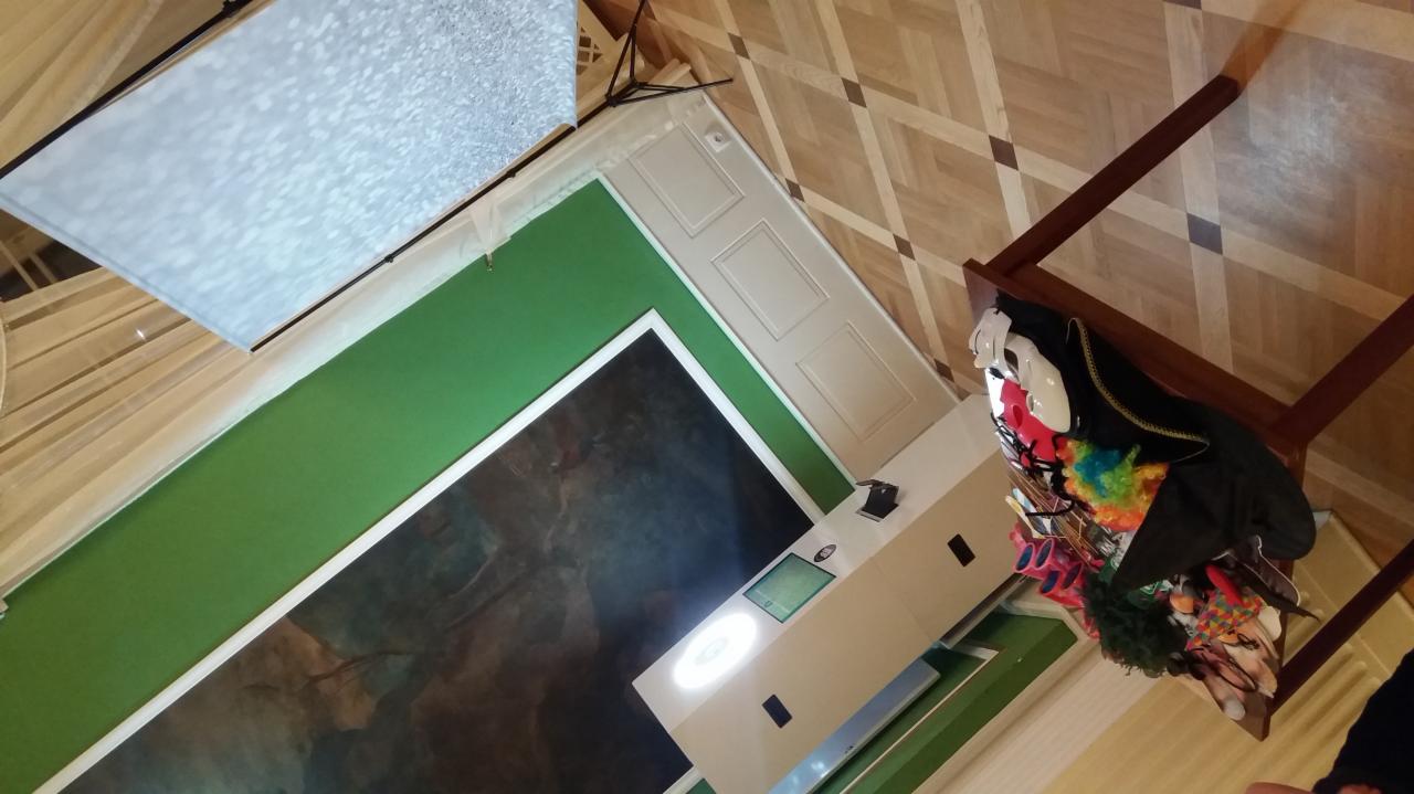 Widok z góry na rozłożoną fotobudkę z rekwizytami, w tym maski i peruki, ustawioną na parkiecie w pomieszczeniu z zielonym tłem i dekoracyjną ścianą.