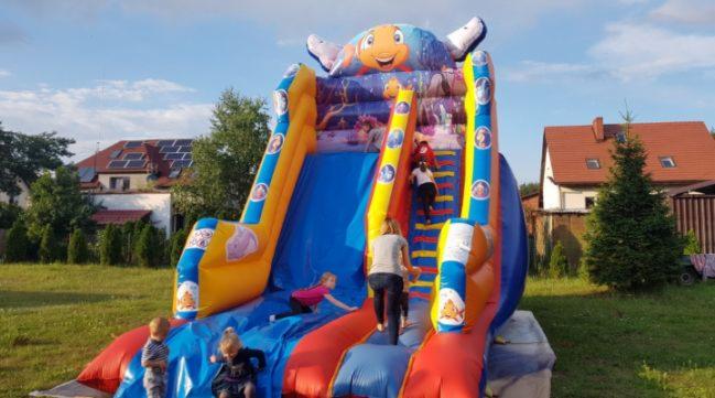 Dmuchana zjeżdżalnia w kształcie ryby z motywami morskimi, dzieci w różnym wieku bawią się na niej w słoneczny dzień na trawniku przed domem z panelami słonecznymi na dachu.