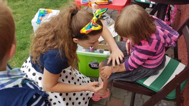 Kobieta w sukience w grochy maluje brokatowy tatuaż na nodze dziecka siedzącego na krześle z zieloną poduszką; w tle zielony pojemnik z napisem 'Party Stars'.