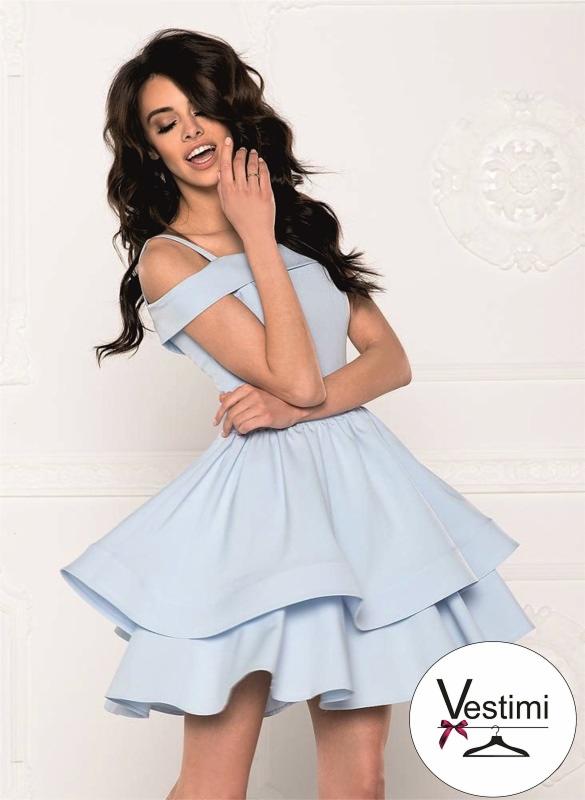 Uśmiechnięta brunetka w błękitnej, dwuwarstwowej sukience z odkrytymi ramionami, pozuje na tle białej ściany ze sztukaterią. W prawym dolnym rogu widoczne logo firmy odzieżowej.