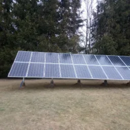 Rząd paneli słonecznych na metalowych podporach, zainstalowany na trawiastym terenie z drzewami iglastymi w tle.