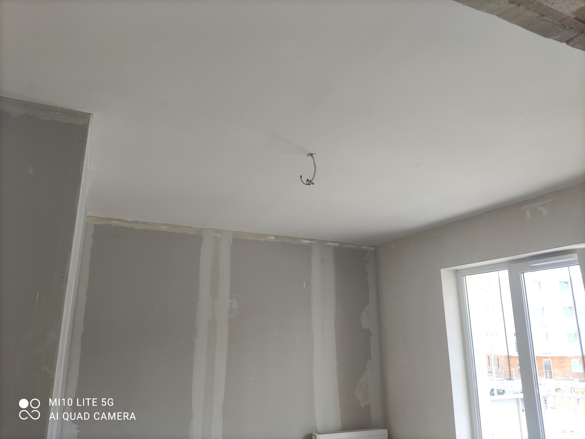 Pokój w trakcie prac wykończeniowych: ściany z płyt gipsowo-kartonowych, częściowo zaszpachlowane, surowy sufit z wystającym kablem, okno z widokiem na budynek.