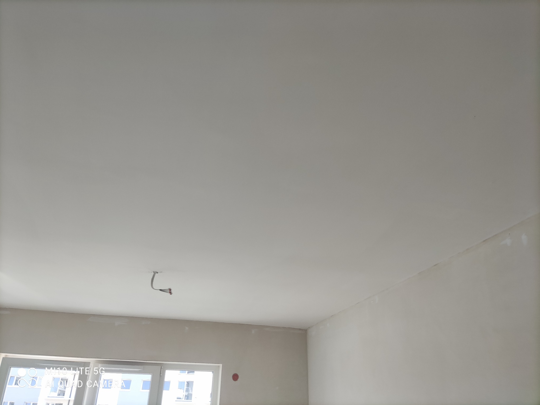 Biały sufit i ściany w trakcie wykańczania, widoczne szpachlowane miejsca, wystające przewody elektryczne i fragment okna z widokiem na budynki