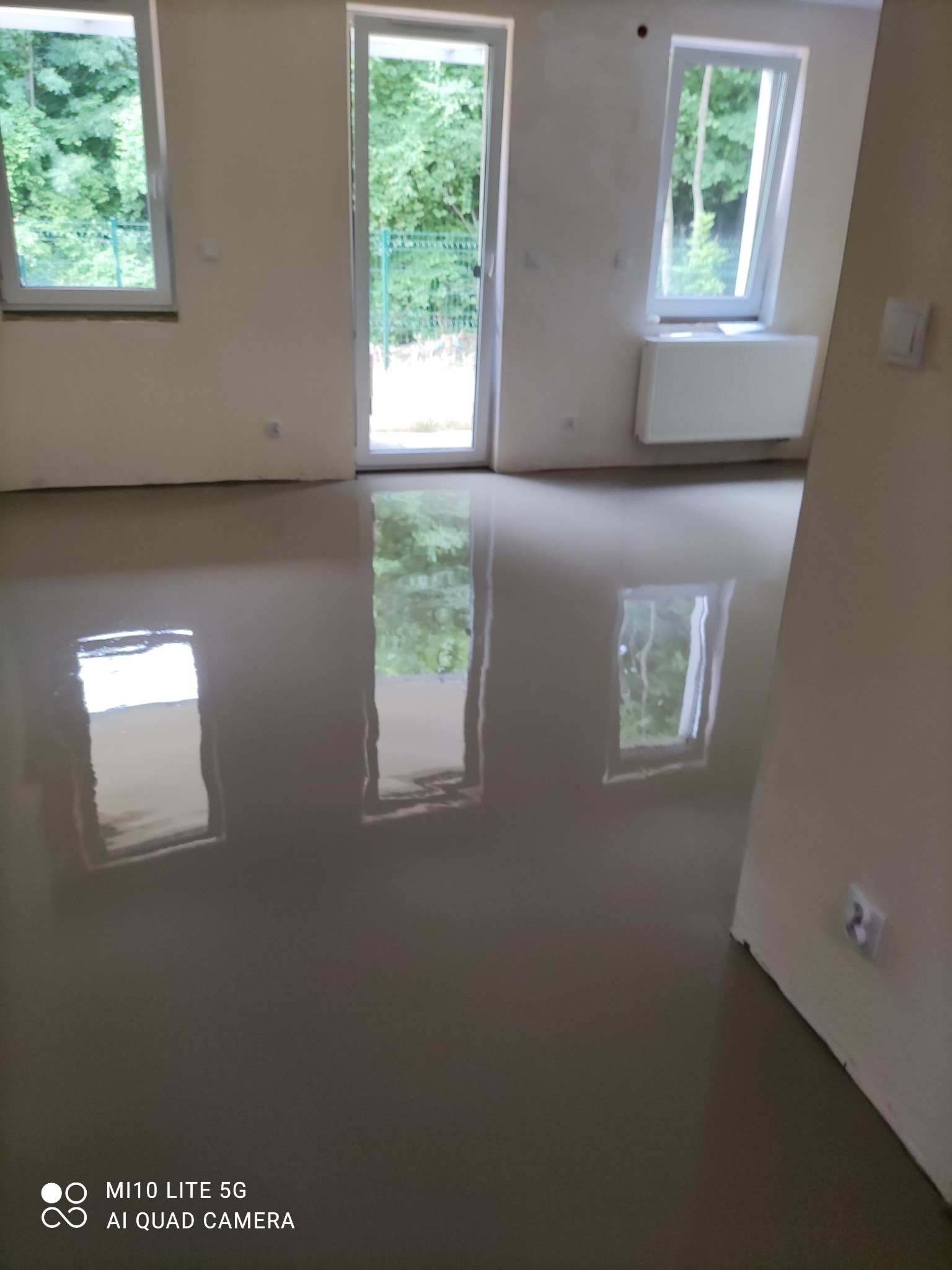 Wylany, mokry podkład podłogowy w jasnym pomieszczeniu z oknami i drzwiami balkonowymi, odbijającymi zieleń drzew na zewnątrz.