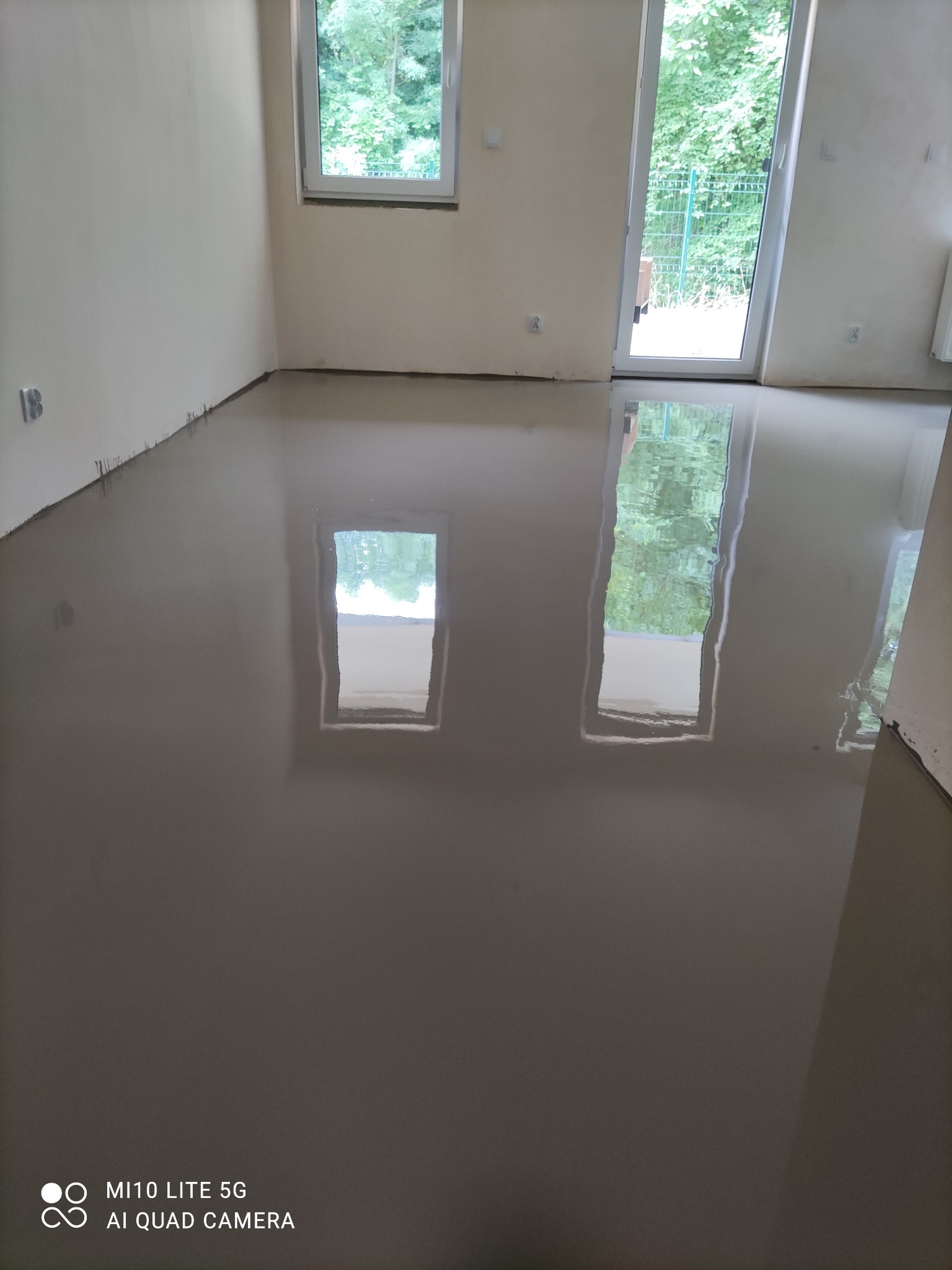 Wylewka samopoziomująca na podłodze w pustym pokoju z oknem i drzwiami balkonowymi, widoczne refleksy światła, ściany przygotowane do malowania.