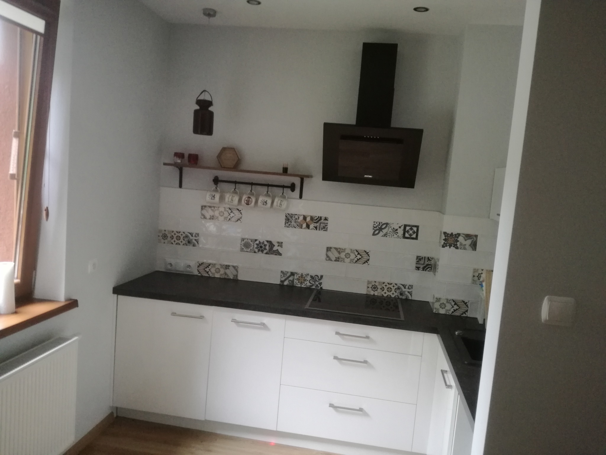 Nowoczesna kuchnia z białymi szafkami, czarnym blatem i dekoracyjnymi płytkami nad blatem, uzupełniona czarnym okapem i półką z kubkami.