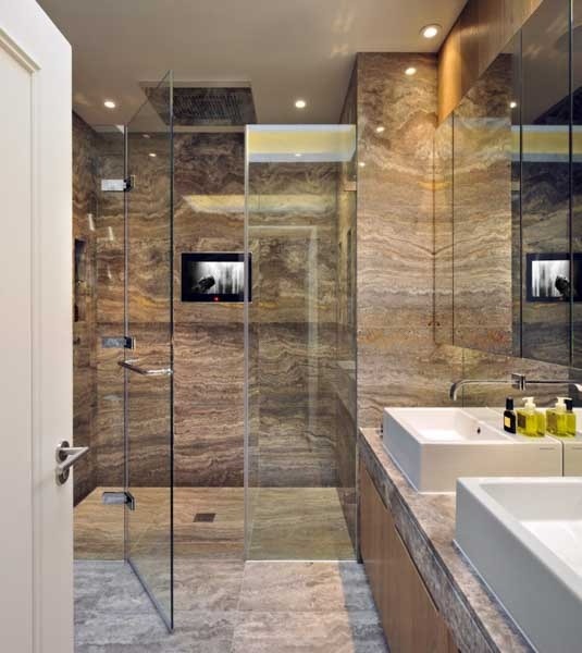 Elegancka łazienka z kabiną prysznicową typu walk-in, wykończona trawertynem w odcieniach brązu, z telewizorem wbudowanym w szklaną ścianę oraz dwiema białymi umywalkami nablatowymi.