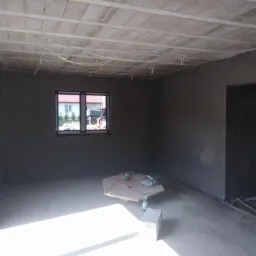 Wnętrze surowego domu po tynkowaniu maszynowym ścian, widoczne przewody elektryczne pod stropem, okno z widokiem na dom, na podłodze narzędzia i buty robocze.