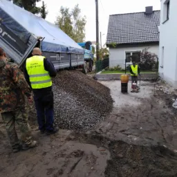 Wywrotka z wysypanym żwirem w Opolu, pracownicy w kamizelkach odblaskowych przygotowują podłoże pod budowę, widoczna zagęszczarka wibracyjna.