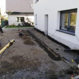 Przygotowanie podłoża pod chodnik z obrzeżami, widoczne narzędzia i materiały budowlane, dom w tle.