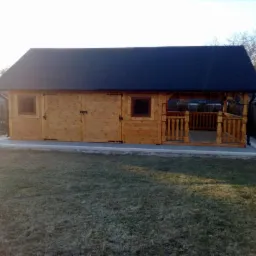 Drewniany domek narzędziowy z zadaszoną werandą, czarny dach, widoczne drzwi dwuskrzydłowe, okno, balustrada werandy, kostka brukowa wokół, trawnik, w tle dom i drzewo.