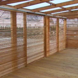 Zadaszona drewniana konstrukcja tarasowa z przesuwnymi panelami żaluzjowymi, zapewniającymi prywatność i ochronę przed słońcem, drewniana podłoga tarasowa, widok z wnętrza konstrukcji.