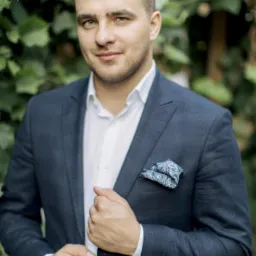 Młody mężczyzna w granatowym garniturze i białej koszuli, z poszetką w niebieskie wzory, pozuje na tle zielonych liści.