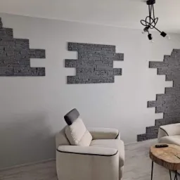 Fragment ściany wewnętrznej z dekoracyjnym, szarym kamieniem ułożonym w geometryczne wzory, uzupełniającym aranżację salonu z beżowymi sofami i drewnianym stolikiem kawowym na tle jasnej ściany...