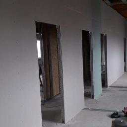 Zakład Remontowo Budowlany - Wnętrze w trakcie montażu ścian działowych z płyty gipsowo-kartonowej, widoczne metalowe profile, surowe drewno, narzędzia na podłodze i fragment okna w tle.