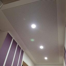 Zakład Remontowo Budowlany - Perforowany, biały sufit podwieszany z wbudowanymi, okrągłymi lampami LED, widok z dołu, ściana z fioletowymi pasami w tle.