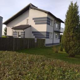 Zakład Remontowo Budowlany - Dom jednorodzinny z jasną elewacją ozdobioną poziomymi szarymi pasami, widoczny z perspektywy ogrodu z niskim żywopłotem i wysokim drzewem iglastym po prawej stronie.