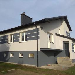 Zakład Remontowo Budowlany - Dwukondygnacyjny dom z jasnoszarą elewacją, ciemnoszarym cokołem i poziomymi pasami. Ciemny komin z płytkami imitującymi łupek. Widoczne rynny i schody wejściowe.