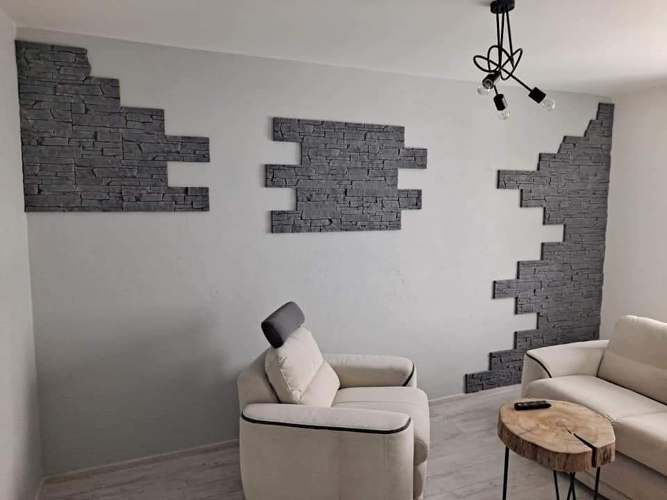 Fragment ściany wewnętrznej z dekoracyjnym, szarym kamieniem ułożonym w geometryczne wzory, uzupełniającym aranżację salonu z beżowymi sofami i drewnianym stolikiem kawowym na tle jasnej ściany...