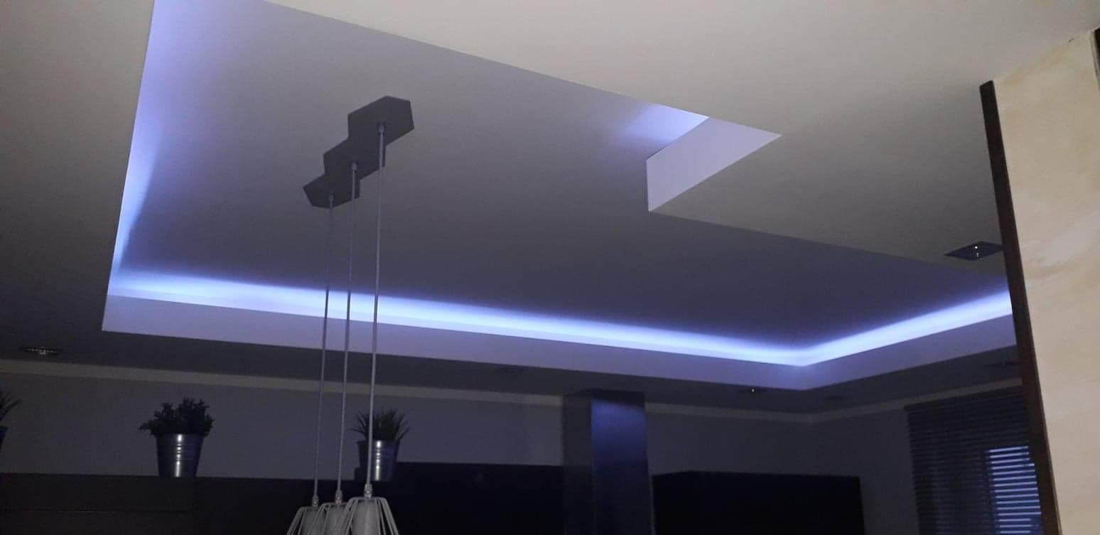 Nowoczesny sufit podwieszany z oświetleniem LED, widoczny fragment lampy wiszącej i rośliny doniczkowej w tle