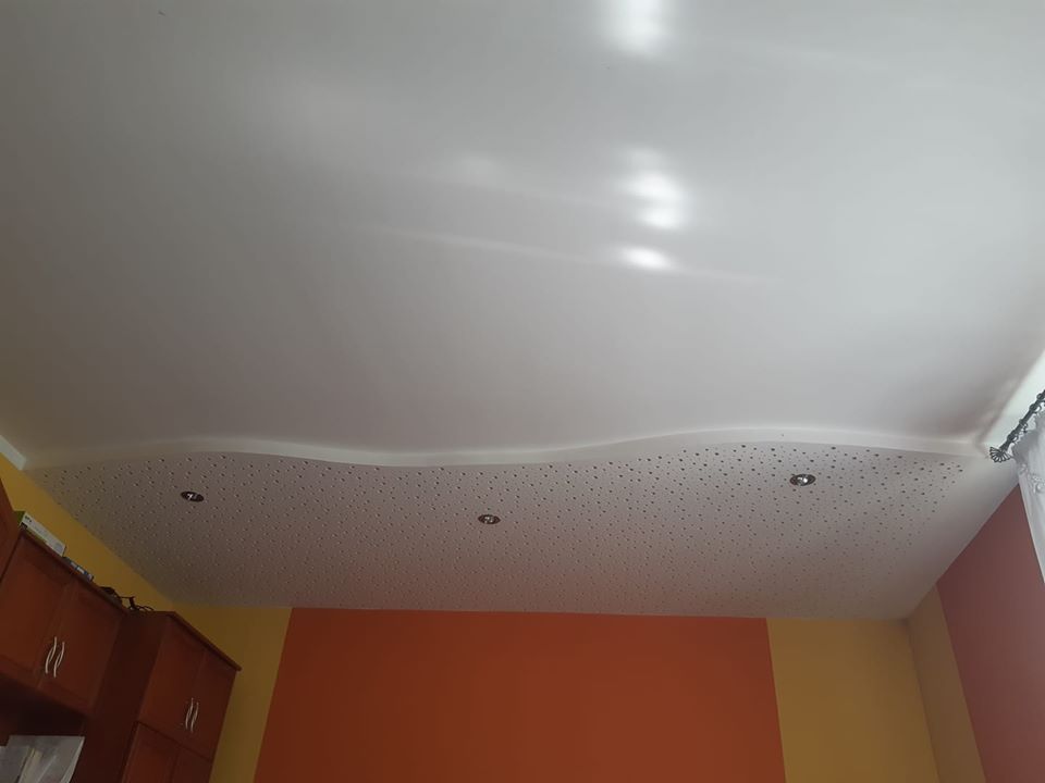 Biały, błyszczący sufit z nieregularnym kształtem i perforowaną częścią z małymi otworami oraz wbudowanymi punktowymi lampami, widok z dołu na pomarańczowo-żółte ściany i fragment szafki.