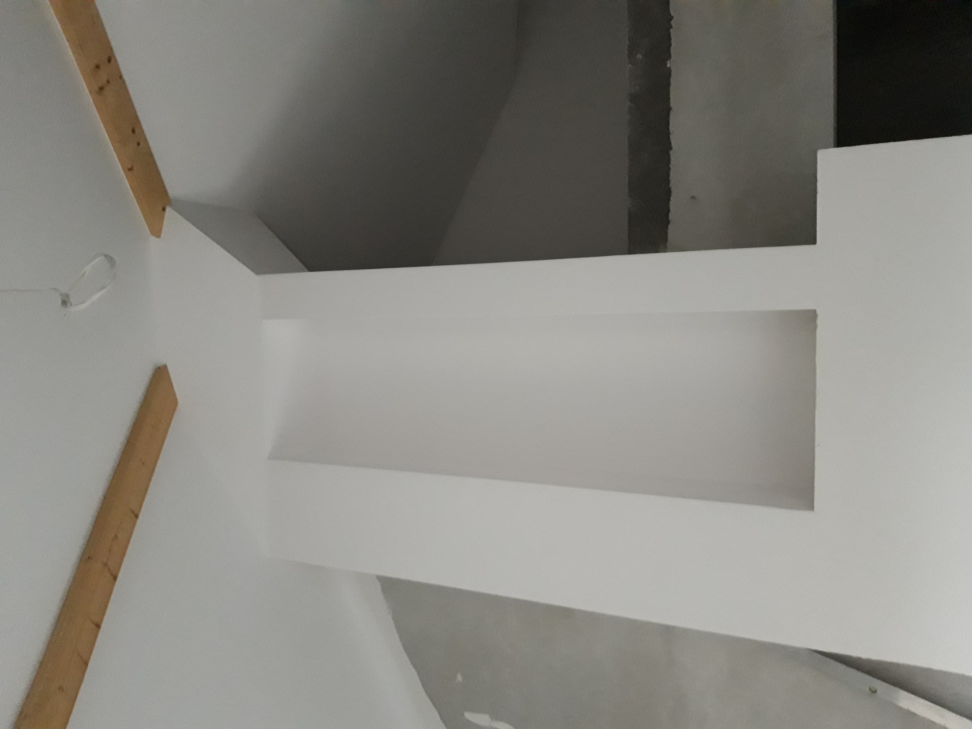 Wnętrze w trakcie remontu, widoczna biała ściana z wnęką, fragment sufitu i drewniane elementy konstrukcyjne, surowe wykończenie.