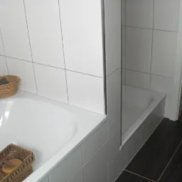 Biała wanna z koszykami wiklinowymi na akcesoria, obłożona białymi płytkami, z widoczną kabiną prysznicową i ciemną podłogą z płytek imitujących drewno.