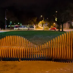 Parametrycznie zaprojektowana ławka drewniana z pionowych desek, o falistym kształcie, ustawiona na tle nocnego krajobrazu miejskiego z zielonym trawnikiem, ulicą i budynkami.