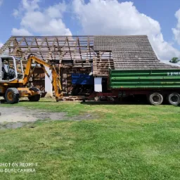 Częściowo rozebrany budynek gospodarczy z odsłoniętą konstrukcją dachu, na tle błękitnego nieba z białymi chmurami, z żółtą koparką Mecalac i przyczepą Lemar w trakcie prac rozbiórkowych.