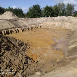 Wyrobisko ziemne z widocznymi śladami po pracy koparki, z niewielkimi zbiornikami wody na dnie i usypaną górą ziemi w tle, częściowo zalesiony teren i budynek w oddali.