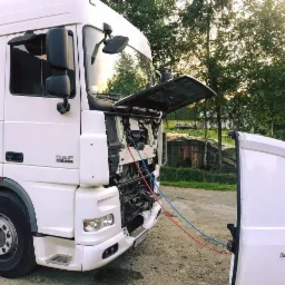 Biała ciężarówka DAF 105.460 z otwartą maską silnika, podłączona przewodami do klimatyzacji z białego busa, na tle drzew i drewnianej szopy.